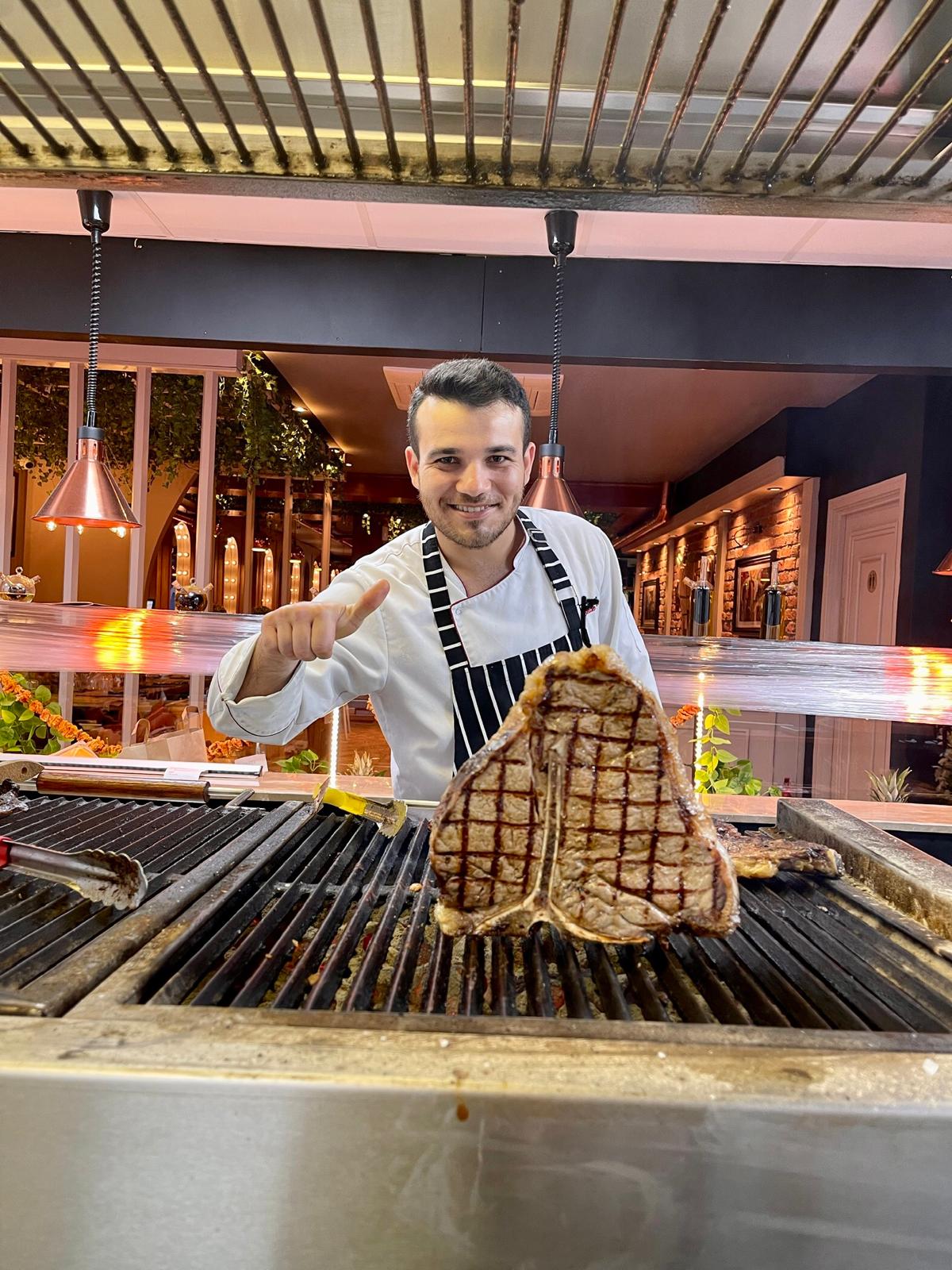 Mikail at the grill holding up a T-bone steak and giving a thumbs up
