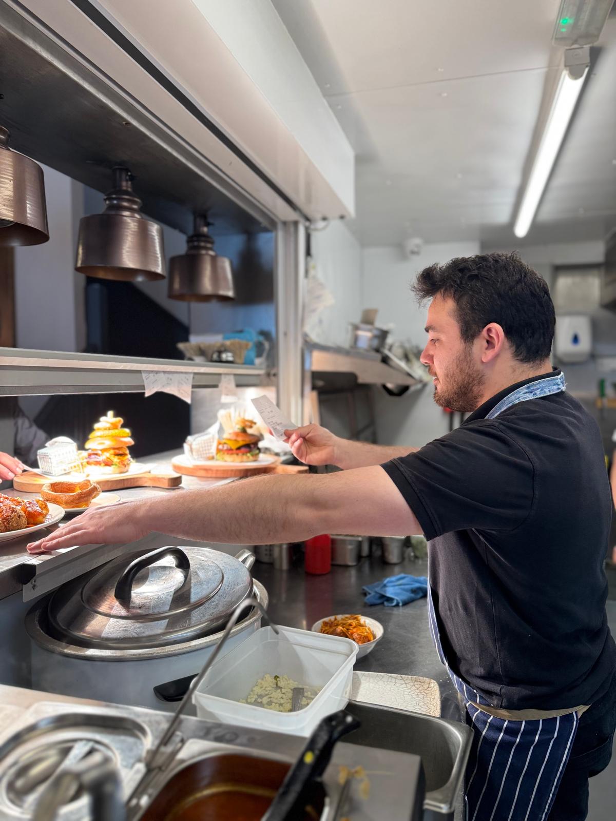 Mikail at the pass reading an order ticket, burgers lined up under the heat lamps